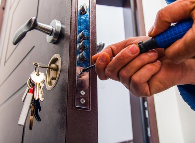 A locksmith using a screwdriver to repair or install a high-security door lock, with a set of house keys hanging from the lock cylinder.