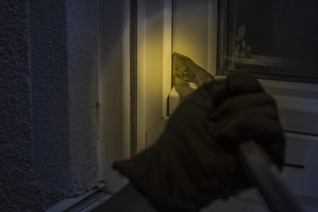 Burglar’s glove covered hands holding a tool to break into a window.